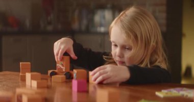 Oyuncu sarışın kız, parlak turuncu oyuncak bloklarıyla dolu masada meşgul. Sevimli çocuk, mutfaktaki evlerle caddeler inşa ediyor. Çocukların boş zamanı. Evin içi güzelmiş. Yüksek kalite 4K görüntü