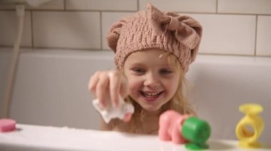 Güzel sarışın kız banyo yapıyor. Sevimli mutlu çocuk duş bonesi takıyor. Bebek banyoda farklı oyuncaklar ve suyla oynuyor. Pediatri konsepti. Eğlence ve rahatlık. Karargah 4k görüntüsü