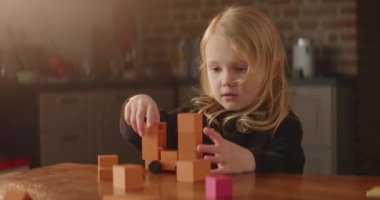 Sarışın kız, portakal rengi tahta oyuncak blokları olan bir masada oynuyordu. Küplerle oynuyordu. Sevimli çocuk tekerlekli bir kule yapıyordu. Çocukların boş zamanı. Aktif hayal gücü. Evin içi güzelmiş. Merkez 4K videosu