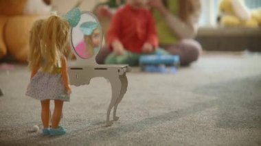Oyuncak bebek oyuncak masa aynasında duruyor, anne ve kızı bebek odası arka planında kuaförlük oynuyorlar, hoş bir iç mekan, yerde oyunlar oynuyorlar. Aile eğlencesi, aktif hayal gücü.