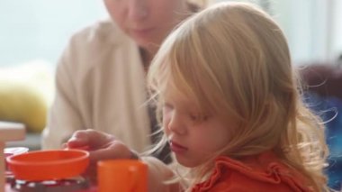 Anne ve kız kahvaltı hazırlama, aile zamanı rol yapma oyunu oynuyorlar. Plastik mutfağı ve oyuncak çaydanlığı olan parlak bir kreş. Hanımlar hayali yiyecek ve içecekler hazırlıyor. Bulaşık ve çatal bıçak. 4k