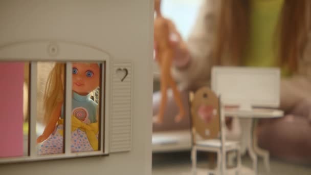 木製のおもちゃの家の窓に立っているブロンドの人形 背景で遊んでいる認識できない10代の女の子 空想的な人工家具 積極的な想像力 家庭での晴れた週末 ロールゲーム 本部4K映像 — ストック動画