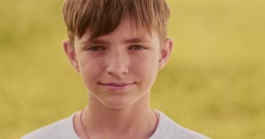 Kameraya bakan genç bir çocuğun yakın portresi. Şehir dışı manzara, rüzgarlı bir gün, sarı tarla, samimi çocuk gülümsemesi, doğal güzellik, Ukraynalı ergen. Yüksek kalite 4k görüntü