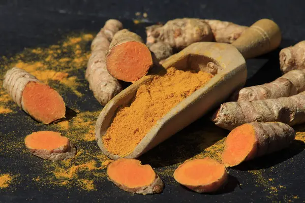 stock image Turmeric powder (Kurkuma) in a wooden spoon and roots on a dark table (curcuma longa)