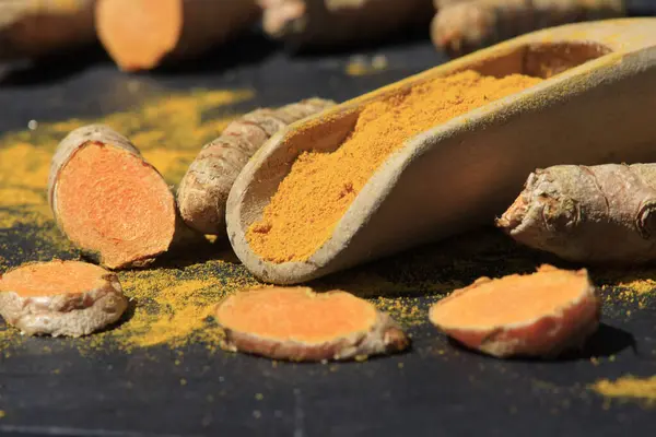 stock image Turmeric powder (Kurkuma) in a wooden spoon and roots on a dark table (curcuma longa)