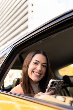 Woman leaning out of the window of a cab in Barcelona looking smiling, holding her cell phone, behind tall office buildings. Attractive and natural caucasian girl with long straight hair.
