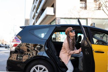 Girl entering yellow and black barcelona cab, with pale pink leather jacket and brown leather cap, smiling woman saying goodbye. In background tall buildings