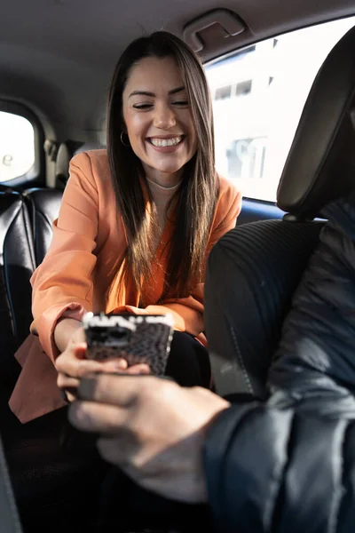 woman inside the back of a car, transport by cab or car with driver, showing her mobile phone to the driver to ask for directions. she is traveling for work with orange blazer and cream sweater, very pretty and smiling.