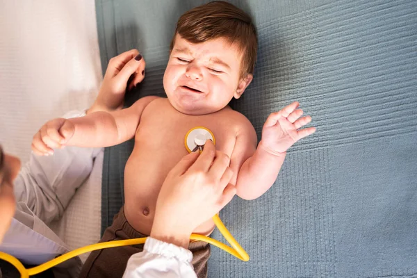 baby crying in bed when doctor auscultates with yellow stethoscope, small blond Caucasian baby