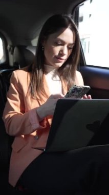 woman inside a car in the back seat, in a cab or car with a driver, writing on her cell phone and working on the computer. Working and doing business on the road