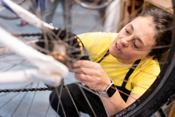 Modern and trendy woman fixing bike gear, girl restoring bike in workshop with bike hanging. on wrist smart watch, black apron and yellow t-shirt.