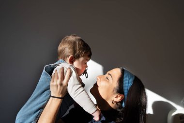 mother alone holding the baby and playing, direct light coming from the window, blue tones and jeans. Latin looking girl, very white baby, blonde hair and blue eyes.