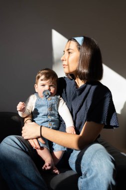single mother sitting on sofa with one year old baby, looking towards window thoughtfully, little one looking at camera smiling and healthy, blue colors and jeans.