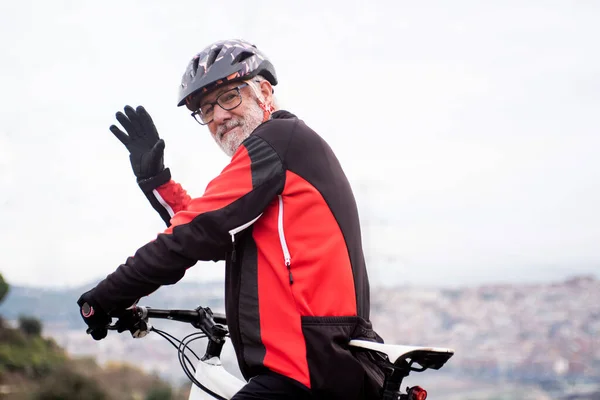 retired elderly male cyclist with bike and equipment waving, city in the background