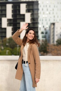 portrait of Caucasian city girl with straight hair waving goodbye, brown cloth coat and jeans, vertical photograph smiling to the side, in the background tall buildings
