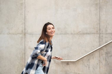 young caucasian girl with straight black hair walking and looking at the camera, casual black and white checkered shirt and jeans, dancing alone in the street with headphones and cell phone. hand in hair and wide smile.