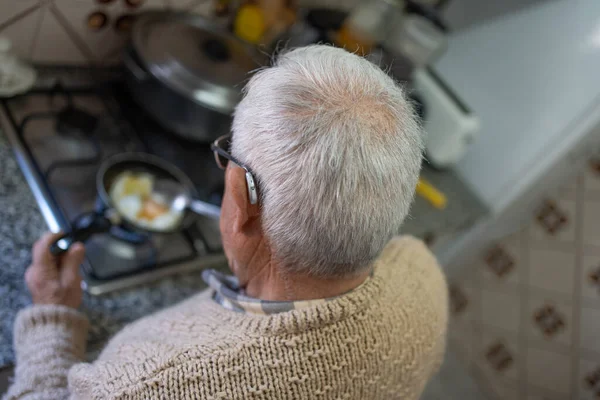 stock image Old man with sonotone cooking fried egg, retired man in brown wool sweater, gray hair, hearing aid preparing breakfast