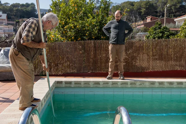 elderly father teaching forty year old son how to clean the bottom of the pool, retired man hand-cleaning a dirty pool, son learning maintenance