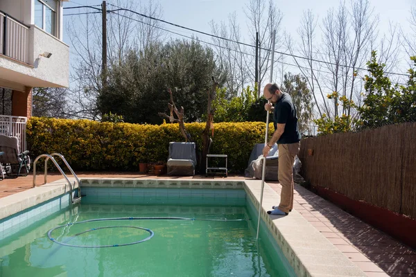 man cleaning swimming pool with manual vacuum cleaner, pool maintenance after winter to prepare the water for summer, private house in urbanization