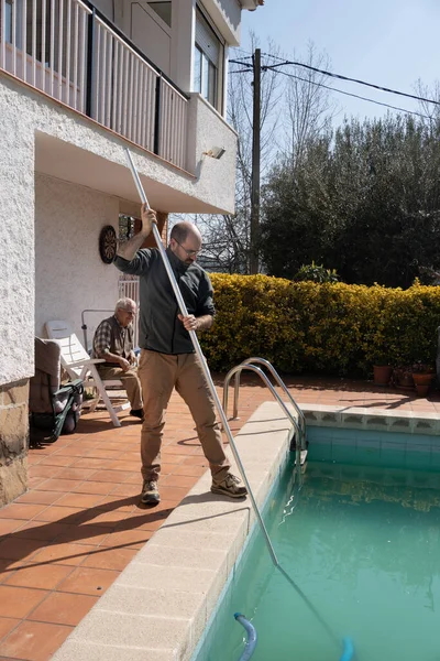 stock image son cleaning the bottom of the pool with the manual pool cleaner, father observing that the pool maintenance is correct, dirty water, preparing the water for the arrival of the summer heat.