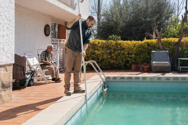 son cleaning the bottom of the pool with the manual pool cleaner, father observing that the pool maintenance is correct, dirty water, preparing the water for the arrival of the summer heat.