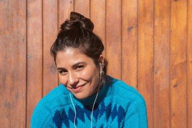 Yüksek topuzlu gülümseyen genç kız portresi, müzik kulaklıklı kadın ve parlak mavi süveter, koyu ahşap arka plan