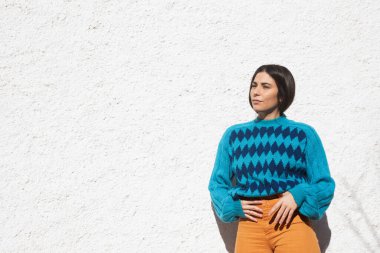 Renkli elbiseli genç Latin kız, doymuş mavi baklava desenli süveter ve turuncu pantolon, dışarıda beyaz duvar arkası, açık güneşli bir gün. Uzayı bir tarafa kopyala