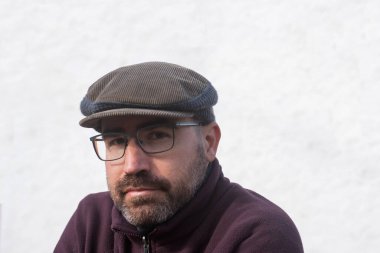 Kahverengi ve gri fitilli kadife ve yün bereli adam. Kırklı yaşlarda, sakallı ve gözlüklü bir adam, kestane rengi bir post takıyor, fotoğraf makinesine bakıyor, açık hava, arka planda bir ev..