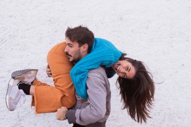 Oğlan patates çuvalı gibi kızı tutuyor, kadın renkli giyiniyor, mavi süveter ve turuncu pantolon giyiyor, beyaz sıva arka plan, açık hava ışığı