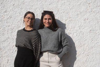 20 'li yaşlarındaki iki kızın kamera önünde portresi. Gri, beyaz ve siyah tonları olan kıyafetler. Beyaz kaplama duvar arkaplanı