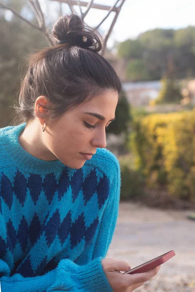 young woman with bow tie looking at cell phone. Outdoors with trees in the background, wearing a blue sweater with diamonds, colorful clothes. natural light with small backlight. Small golden earrings in the ears