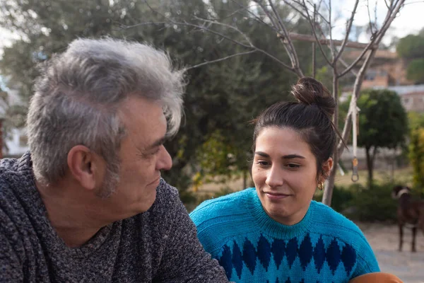 Baba ve kızı dışarıda ciddi bir ifadeyle konuşuyorlar. Gri saçlı, gri kazaklı, yirmili yaşlarda papyonlu ve doymuş mavi kazaklı bir kız. arkaplan ağaçlarında.