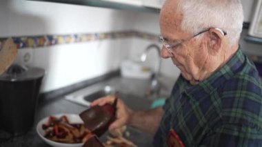 Yaşlı adam büyük kırmızı inek biberi kesiyor, yaşlı adam evde tek başına yemek pişiriyor, seksen yaşındaki baba kızarttığı için biber kesiyor.