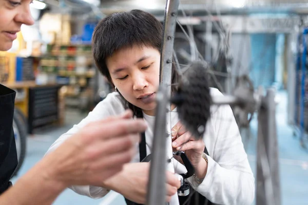 Caucasian woman explaining bicycle wheel alignment to Asian girl in bike shop. Women learning to fix bicycles and forming a multi-ethnic team.
