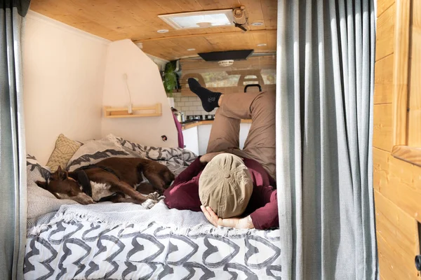 camper van seen from the back, rear doors open where we see the furniture and bed, inside a man and a dog sleeping. outside two camping chairs.