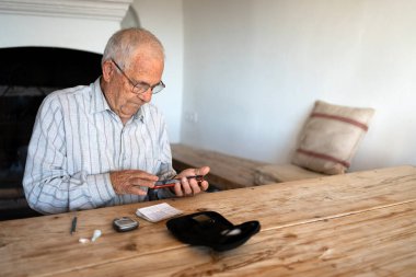 80 yaşında bir adam kan şekeri seviyesini ölçüyor ve sağlığını korumak için kağıt ve cep telefonuna yazıyor. Banklar ve ahşap masa ile kırsal alan..