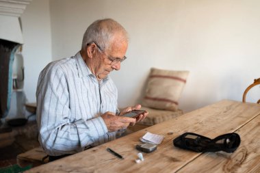 80 yaşında bir adam kan şekeri seviyesini ölçüyor ve sağlığını korumak için kağıt ve cep telefonuna yazıyor. Banklar ve ahşap masa ile kırsal alan..