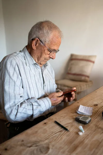 80 yaşında bir adam kan şekeri seviyesini ölçüyor ve sağlığını korumak için kağıt ve cep telefonuna yazıyor. Banklar ve ahşap masa ile kırsal alan..