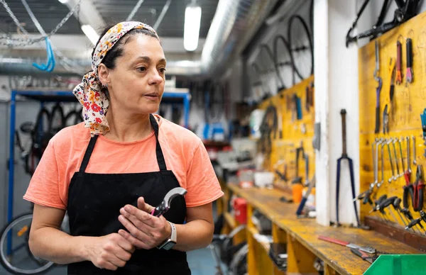 woman with tool in hand looking for another on wall full of tools, female mechanic in black apron, T-shirt and headscarf in her forties.