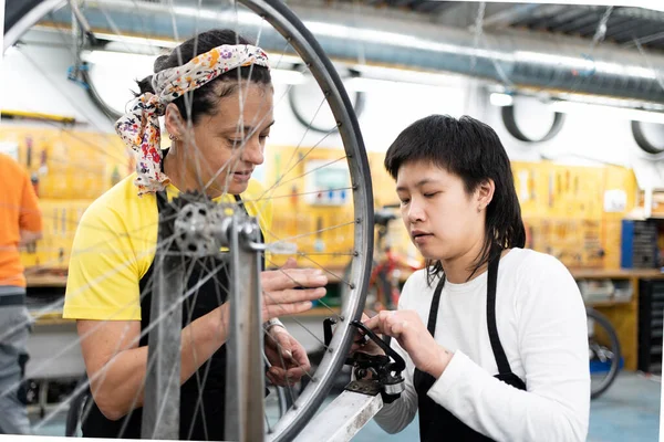 female bicycle mechanic teaching young asian girl to align bike wheel in a self-repair shop. two girls in black aprons to fix bikes.