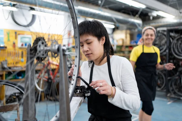 young asian woman in collective self-repair workshop alienating bicycle wheel with tool. Dressed in black apron, another female mechanic out of focus behind.