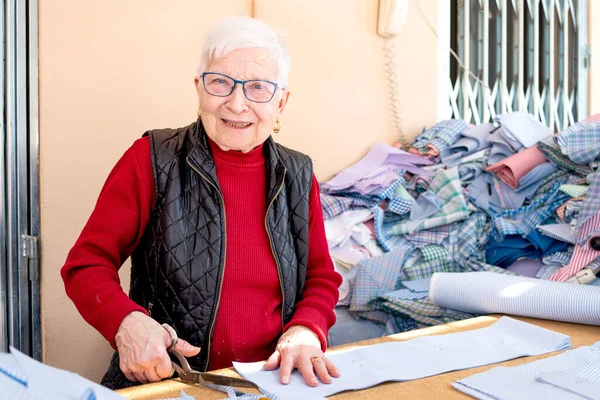 woman cutter in her eighties posing for the camera, active seamstress cutting striped fabric typical of traditional gowns, short white hair and smiling.