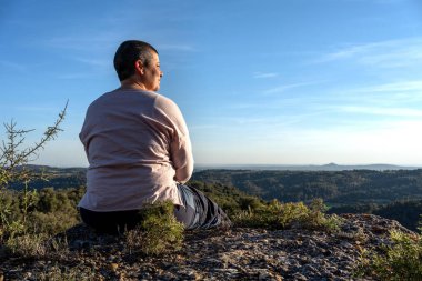Sırtı geniş bir günbatımı manzarasında dalgın, kısa saçlı, biraz şişman, yansıtan ve manzaranın tadını çıkaran ikili olmayan biri..