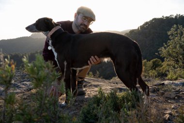 Bir kayanın tepesinde bereli bir adam gün batımında küçük bir tepenin üstünde doğa manzaralı bir köpeği okşuyor. Köpek uzağa bakıyor.