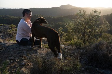 Günbatımında, doğanın ortasında bir köpekle, bir köpek kayasının tepesinde ve çok kısa saçlı, geniş bir orman ve tarla manzarası olan bir kadınla, ikili olmayan bir insan..