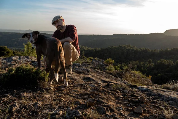 man in beret on top of a rock petting a dog in the sunset, on top of a small hill with great views of nature. Dog and man looking at camera