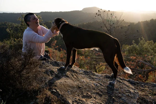 non-binary person in the sunset with a dog in the middle of nature, on top of a dog rock and a woman with very short hair with a wide landscape of woods and fields in the background.