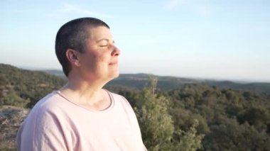 Gün batımında gözleri kapalı meditasyon yapan bir kadın, çok kısa saçlı, açık pembe uzun kollu tişörtlü bir kadın.