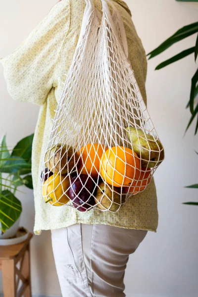 stock image unrecognisable woman with reusable net bag full of fruit, plant environment and smooth wall. Shirt in yellow tones.