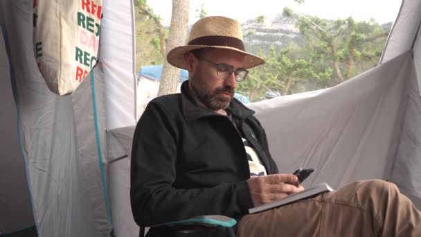 戴着探险家帽子的男人看着他的手机 在露营者的帐篷里舒服地读着书 在暴风雨开始的时候 外面刮起了一阵大风 充满了冒险精神 — 图库视频影像